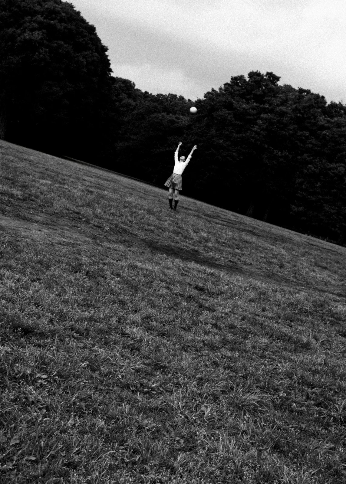 Black and white photo of a young girl in a plaid skirt playfully tossing a ball into the sky on a sloped grassy field, surrounded by dense trees. The fleeting moment captures the essence of mono no aware, highlighting the impermanence and ephemeral joy of childhood.